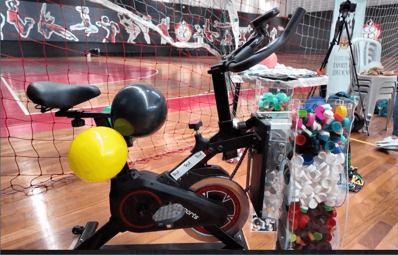 Bike Recicladora com reservatórios de tampinhas em quadra esportiva durante o evento IEE